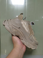 Side view of a light beige Balenciaga Triple S sneaker showing layered panels, mesh sections, branded side detailing, and a chunky textured sole.