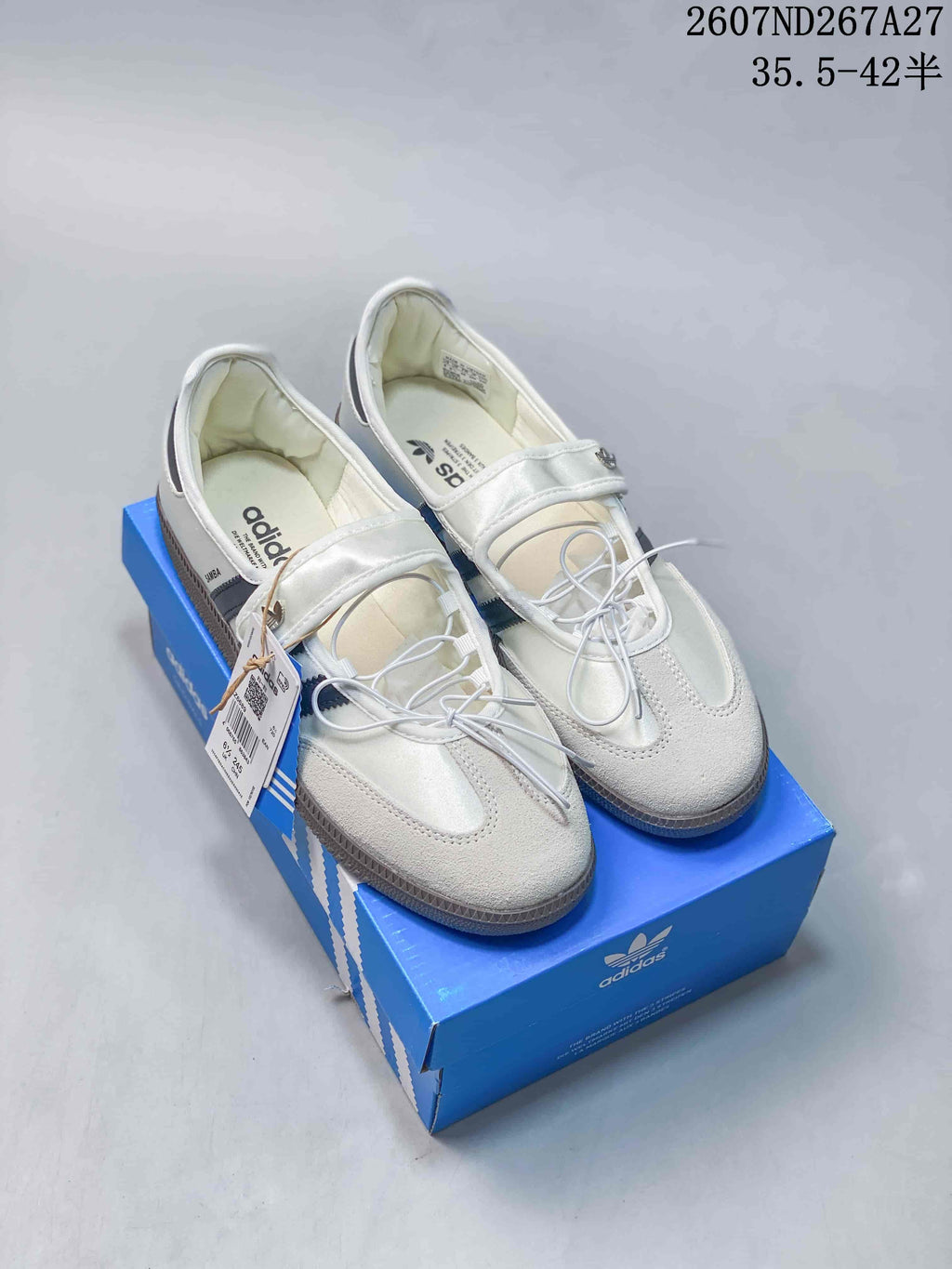Pair of white adidas Samba Jane sneakers displayed on a blue adidas shoebox, showing the strap, laces and gum-colored outsole.