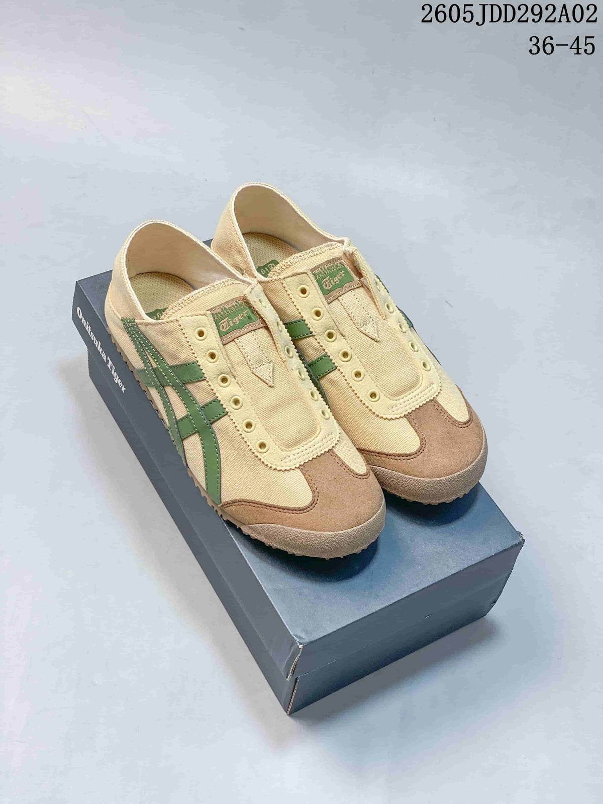 Pair of beige Onitsuka Tiger slip-on sneakers with green stripes and brown toe overlays displayed on a branded box; image text shows style code 2605JDD292A02 and size range 36–45.