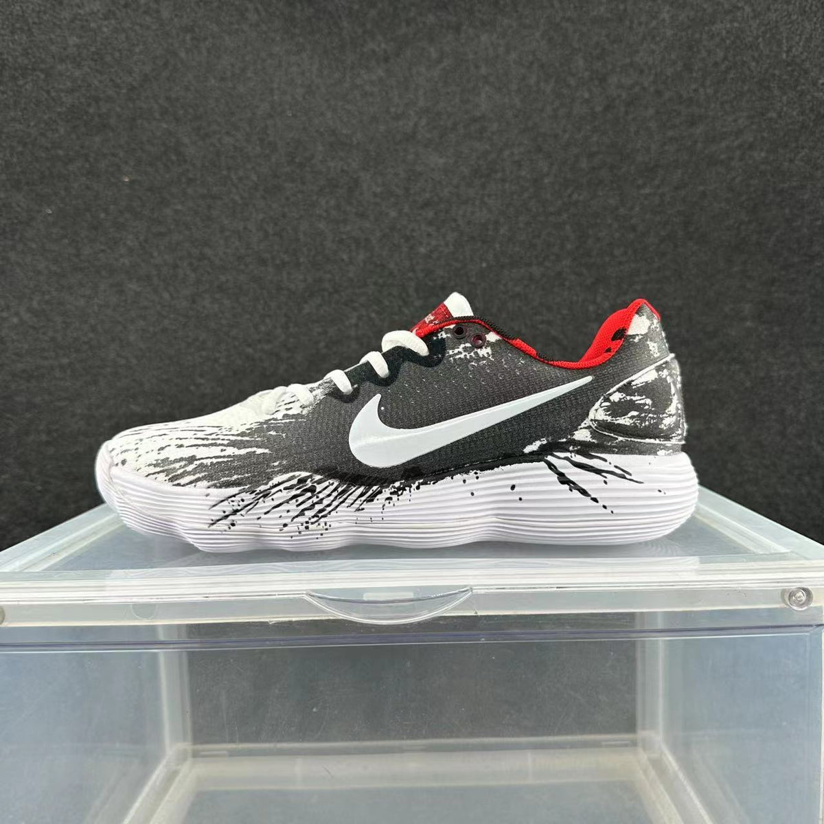 Side view of a white, black, and red Nike low-top sneaker with a white swoosh-style logo and black splatter details on the sole.