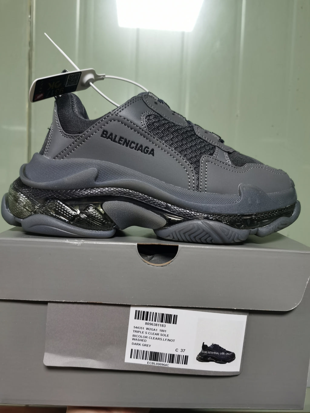 Side view of the dark grey sneaker resting on its box, with the box label showing Triple S Clear Sole, style code 544351 W2GA1 1801, color Dark Grey and sample size C 37.