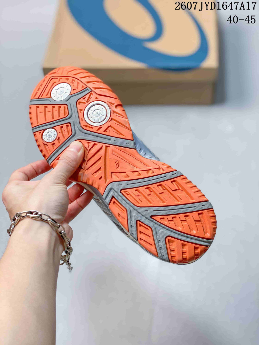 Close-up of the orange and grey patterned outsole on an ASICS tennis shoe; style code 2607JYD1647A17 and size range 40–45 are visible.