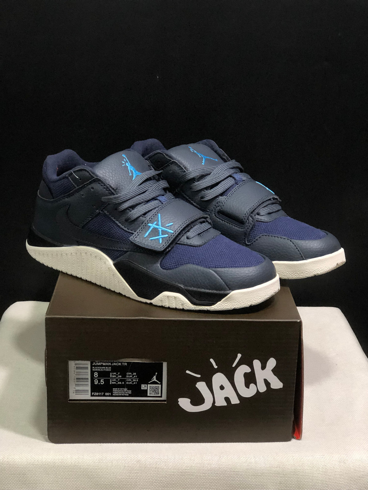 Pair of dark black and navy Jordan Jumpman Jack sneakers displayed on a branded box, showing blue details and cream outsoles.