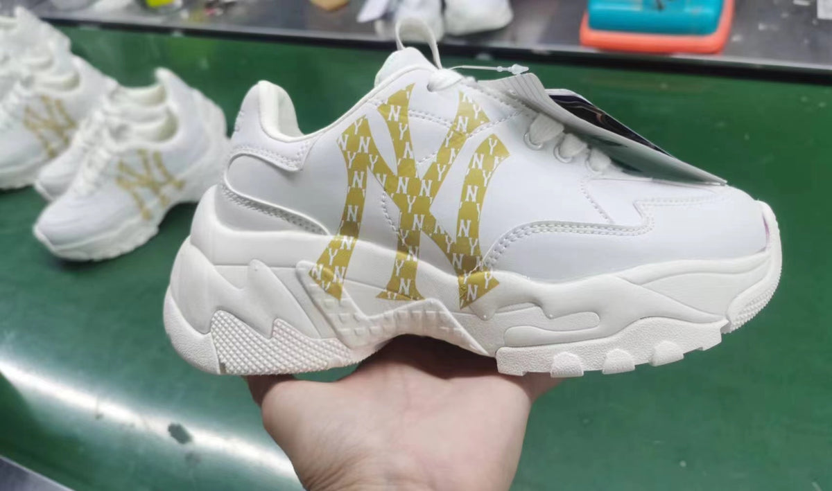 White chunky low-top sneaker with a repeated gold NY-style pattern on the side, white laces, and a sculpted outsole.