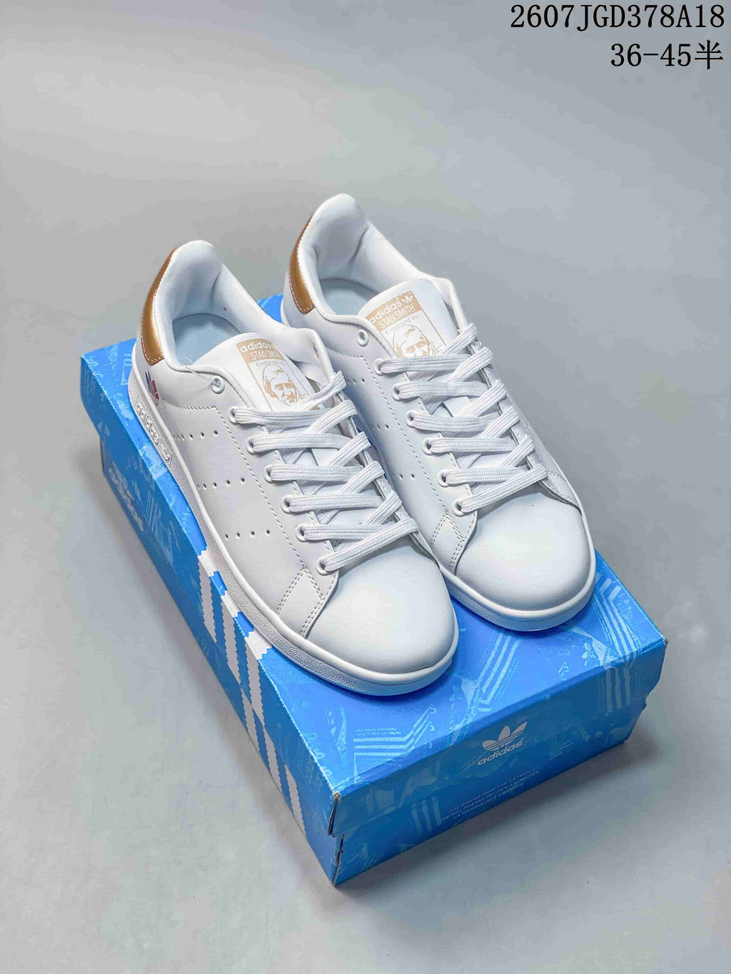 Pair of white adidas Stan Smith sneakers on a blue shoebox, shown with gold-colored heel accents and tongue details.