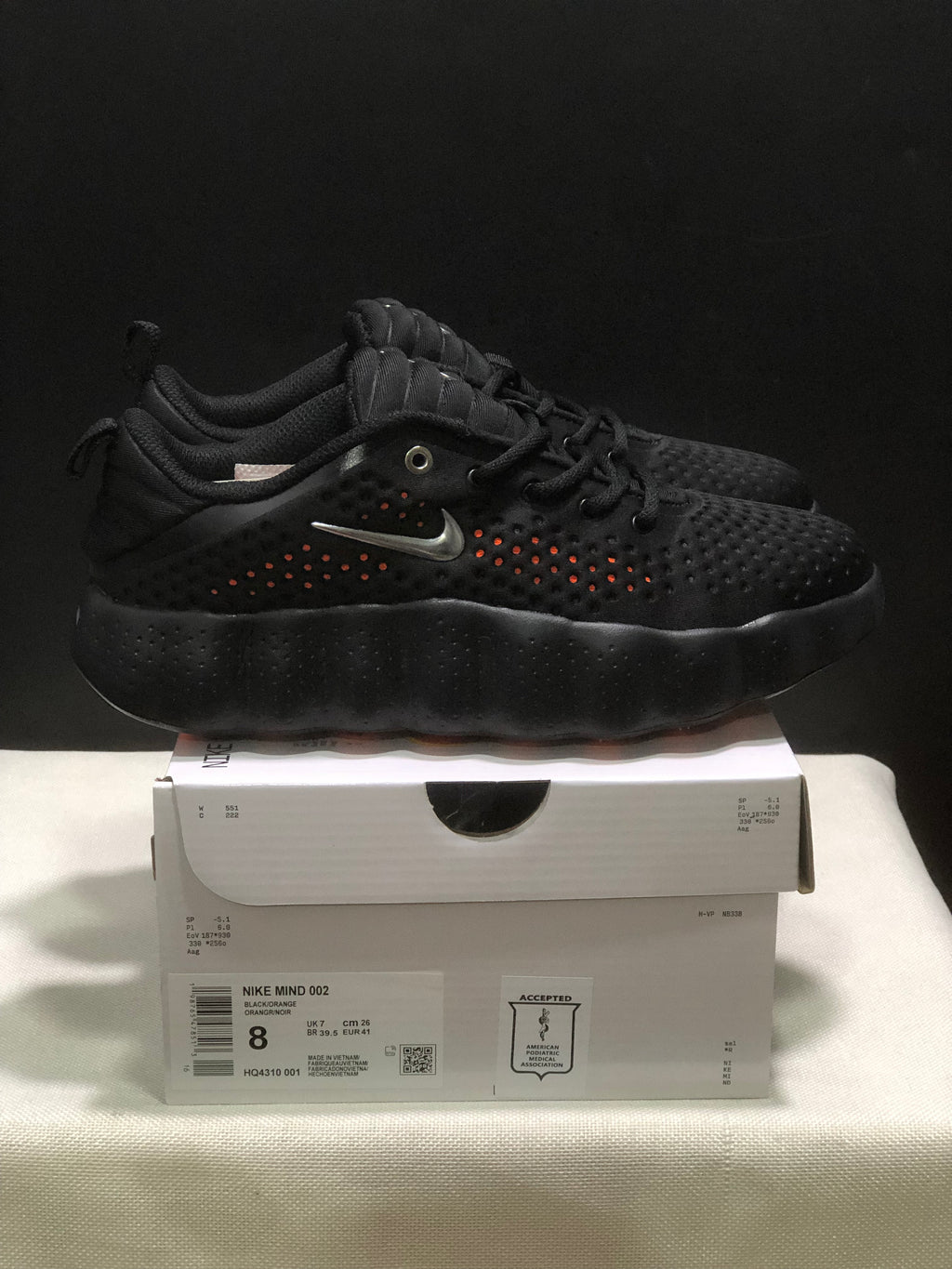 Side view of black Nike Mind 002 sneakers displayed on a shoebox, showing orange perforation accents and the box label.
