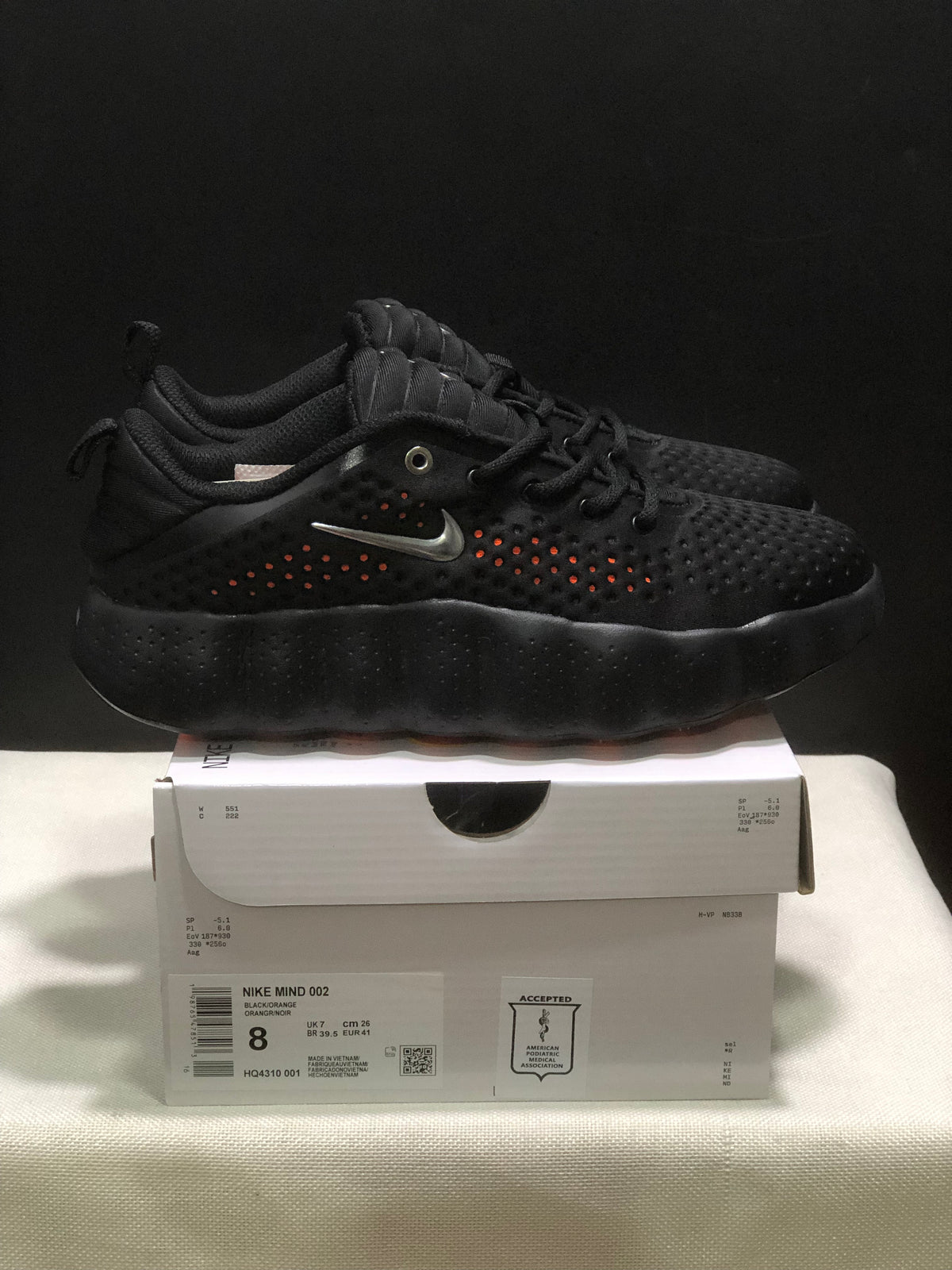 Side view of black Nike Mind 002 sneakers displayed on a shoebox, showing orange perforation accents and the box label.