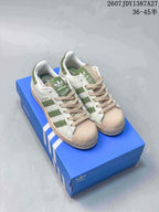 Pair of white adidas Superstar sneakers with green striped detailing, beige shell toes, beige laces, and a blue adidas shoebox; image text shows JDY1387A27 and sizes 36–45.