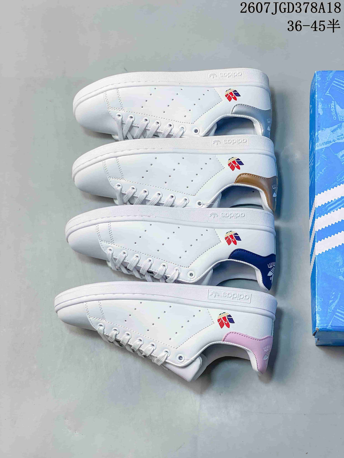 Four white adidas Stan Smith sneakers arranged side by side, showing navy, gold, and pink heel accents alongside a blue shoebox.