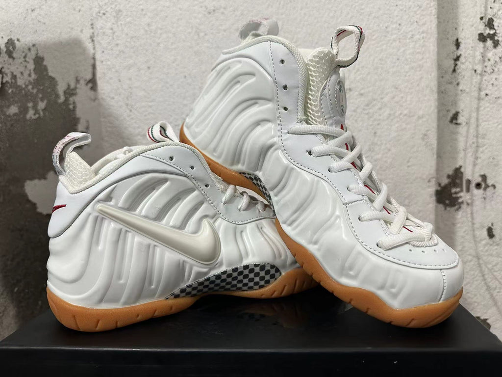 Pair of white Nike Foamposite sneakers shown from the side with sculpted panels, Swooshes, checkerboard accents, and gum outsoles