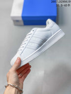 Side profile of a white Adidas Superstar sneaker showing the three-stripe panel, perforations and low-top silhouette.
