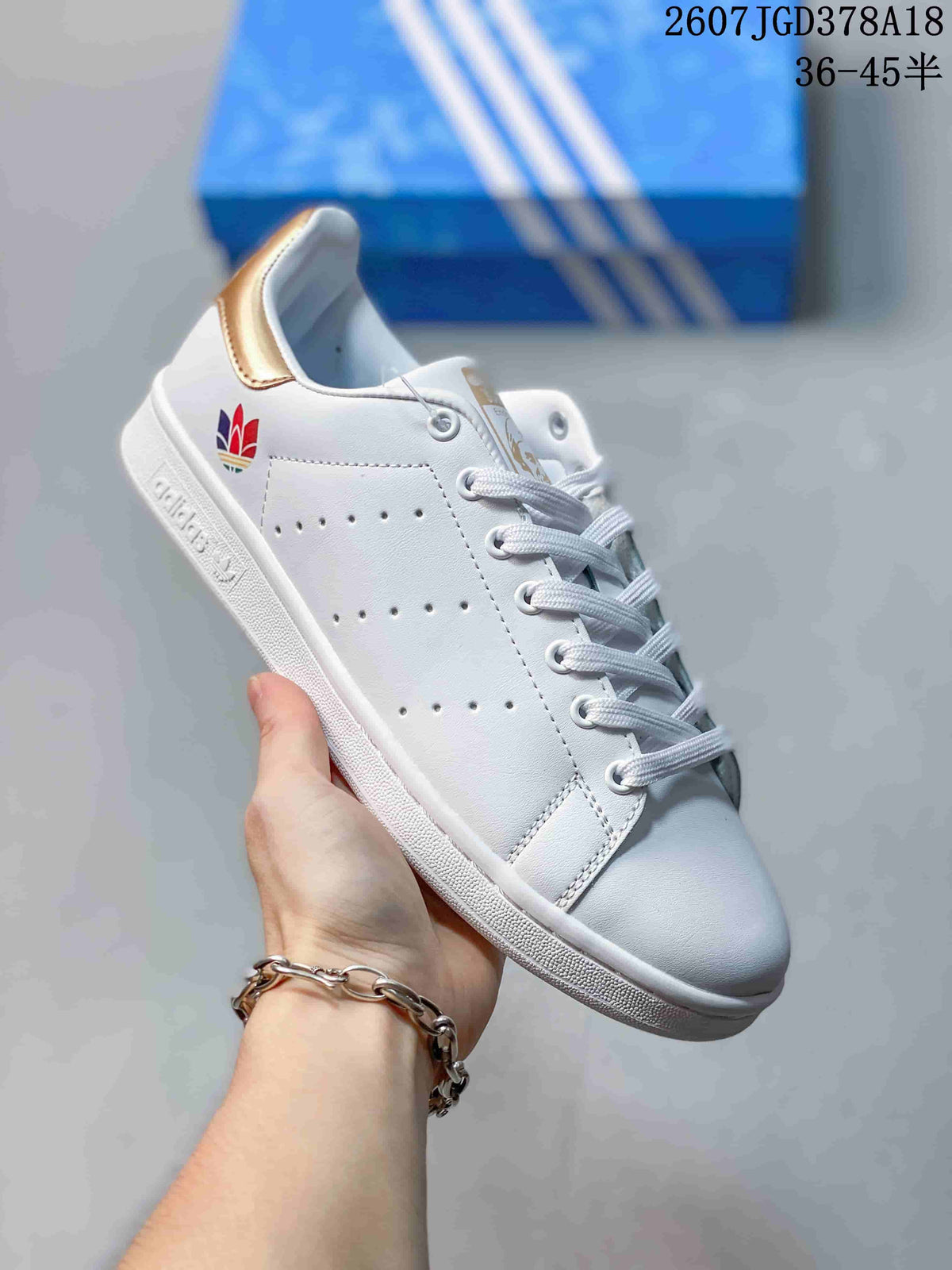 Close side view of a white adidas Stan Smith sneaker with a gold-colored heel accent, perforated upper, and lace-up front.