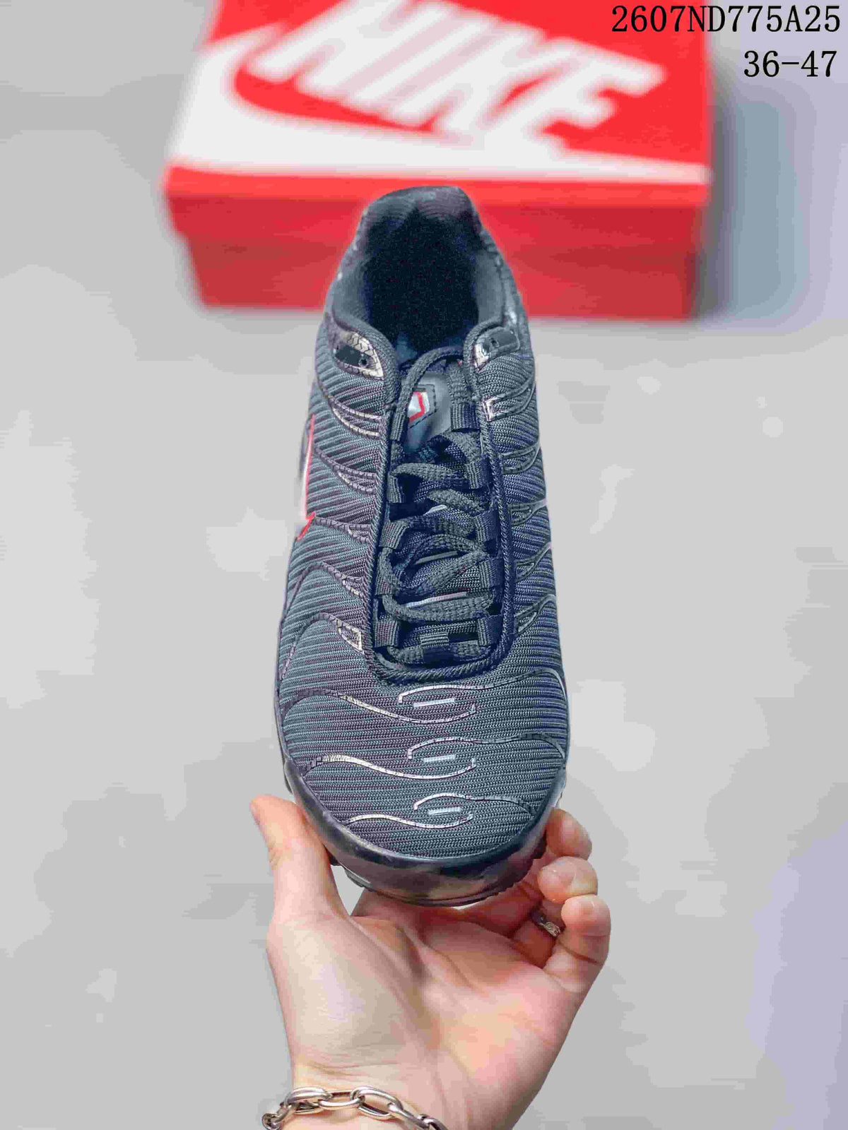 Top view of a black Nike Air Max Plus TN sneaker showing the lace-up design, textured upper, and silver detailing; overlay text shows code 2607ND775A25 and size range 36–47.