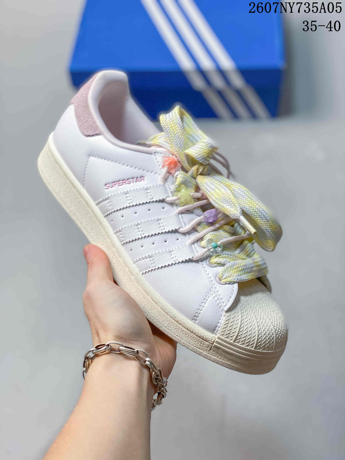 Angled side view of the white Adidas Superstar sneaker with pink accents, pastel laces, and a cream-colored sole.