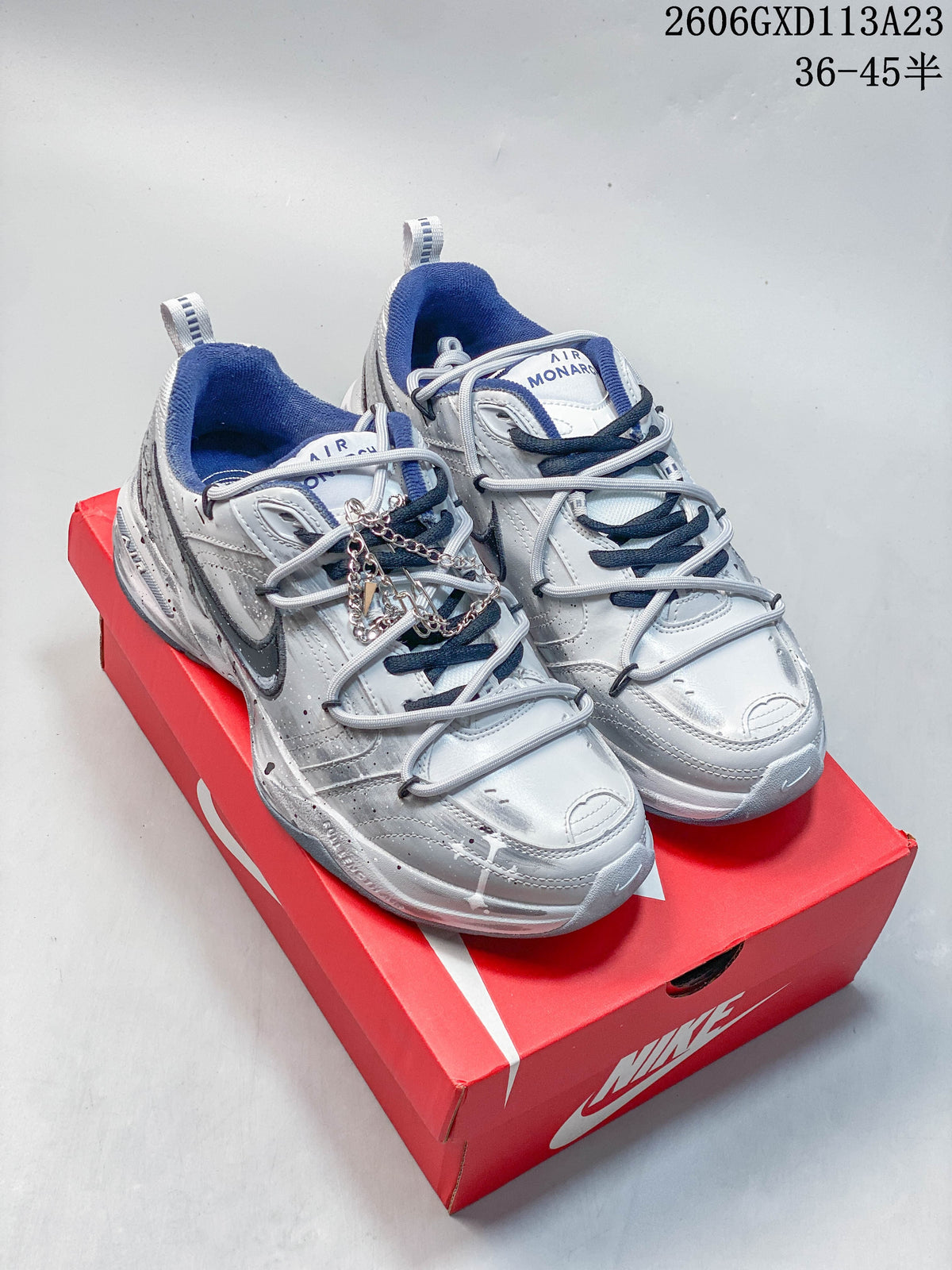 Pair of silver Nike Air Monarch IV Tekno sneakers with navy accents displayed on a red shoebox.