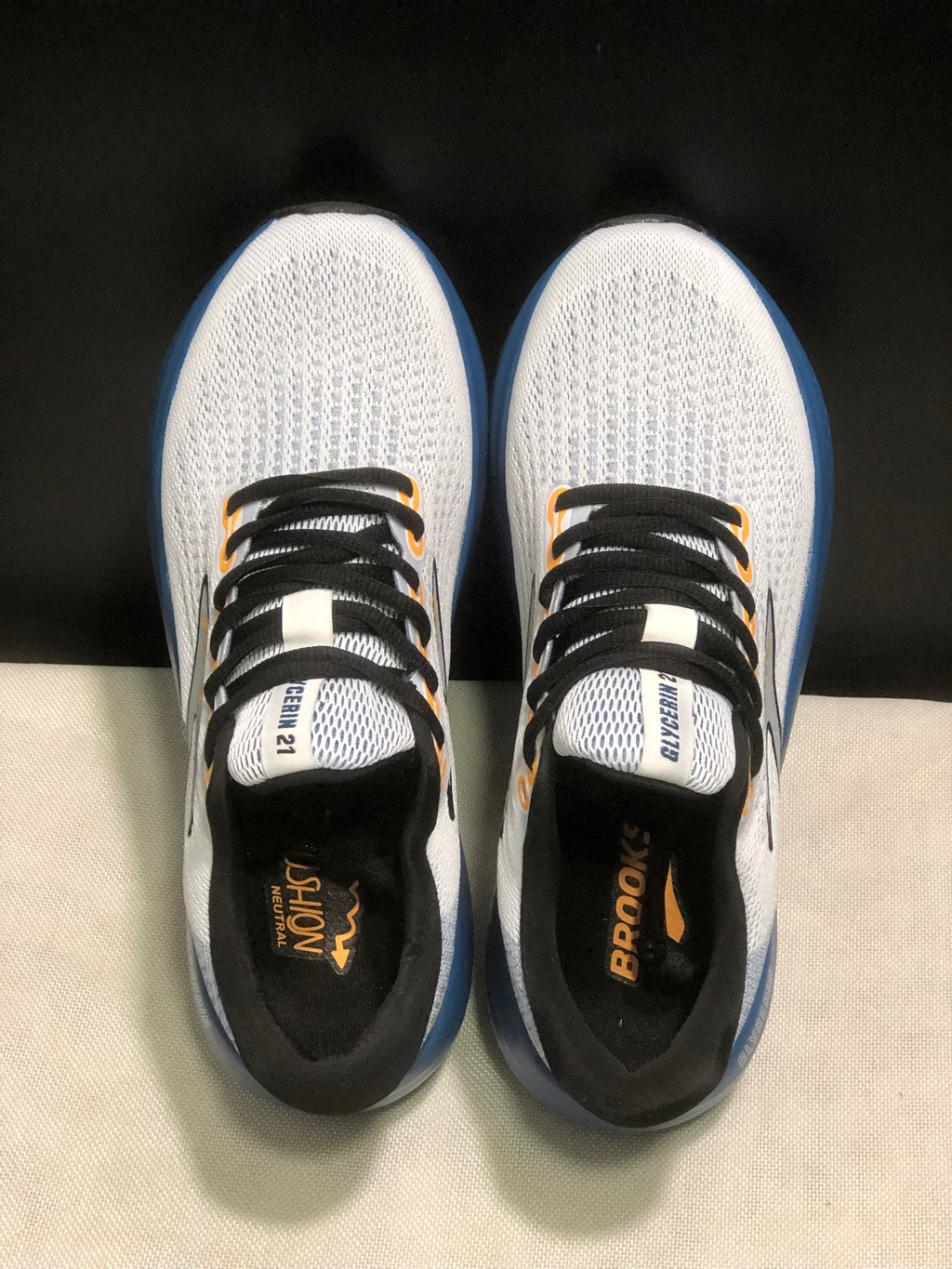 Top-down view of the pair showing the white mesh-like uppers, black laces, orange eyelet accents, and branded insoles.