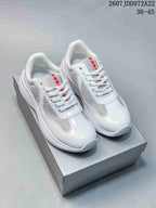 Pair of white Prada America's Cup low-top lace-up sneakers displayed on a shoebox, with gray mesh-style panels and red tongue accents.