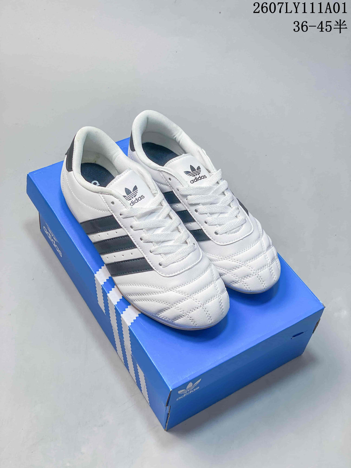 Pair of white Adidas Taekwondo low-top shoes with dark stripes and gum-colored soles, shown on a blue box.