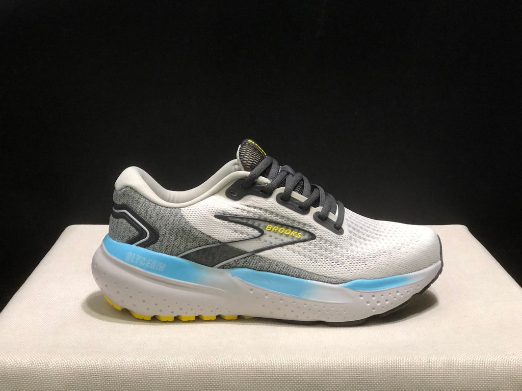 Side profile of one Brooks Glycerin 21 shoe against a dark background, showing its white and grey colorway, black laces, blue midsole accent, and yellow outsole details.