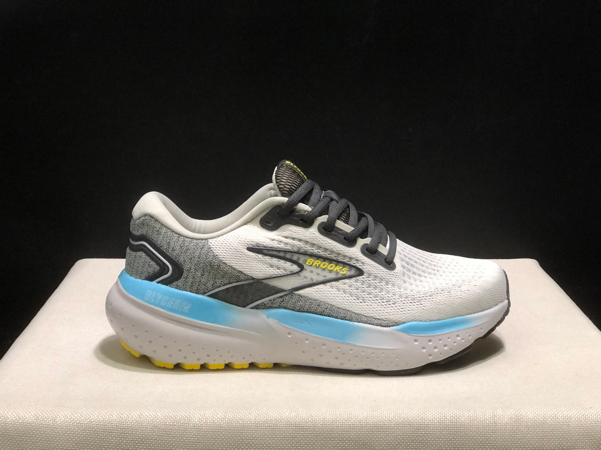 Side profile of one Brooks Glycerin 21 shoe against a dark background, showing its white and grey colorway, black laces, blue midsole accent, and yellow outsole details.
