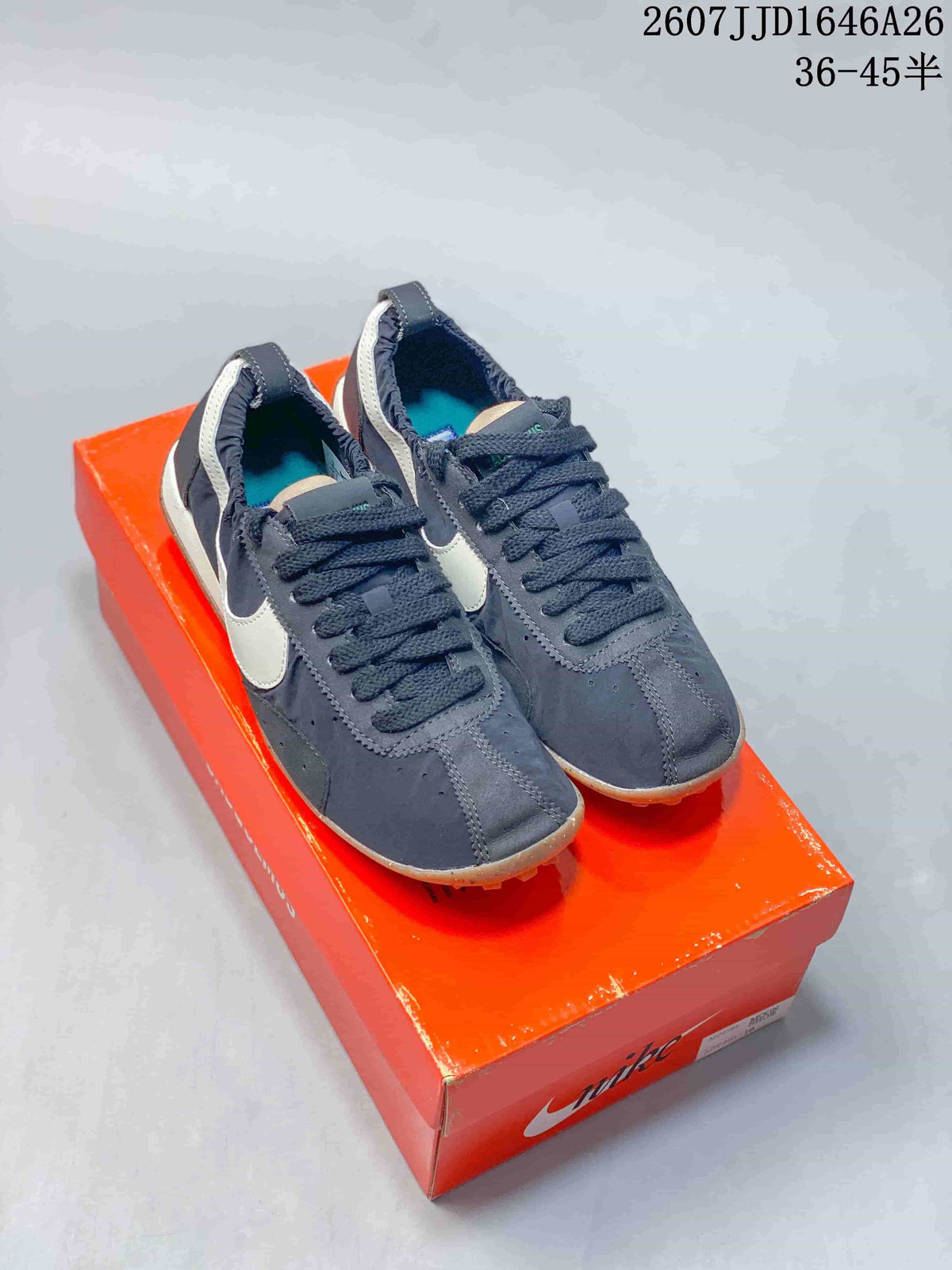 Pair of dark Nike x Jacquemus low-top sneakers with white Swooshes displayed on an orange shoebox.
