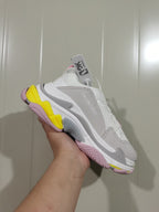 Angled view of the white Balenciaga Triple S sneaker showing layered panels, grey detailing and pink and yellow outsole sections.