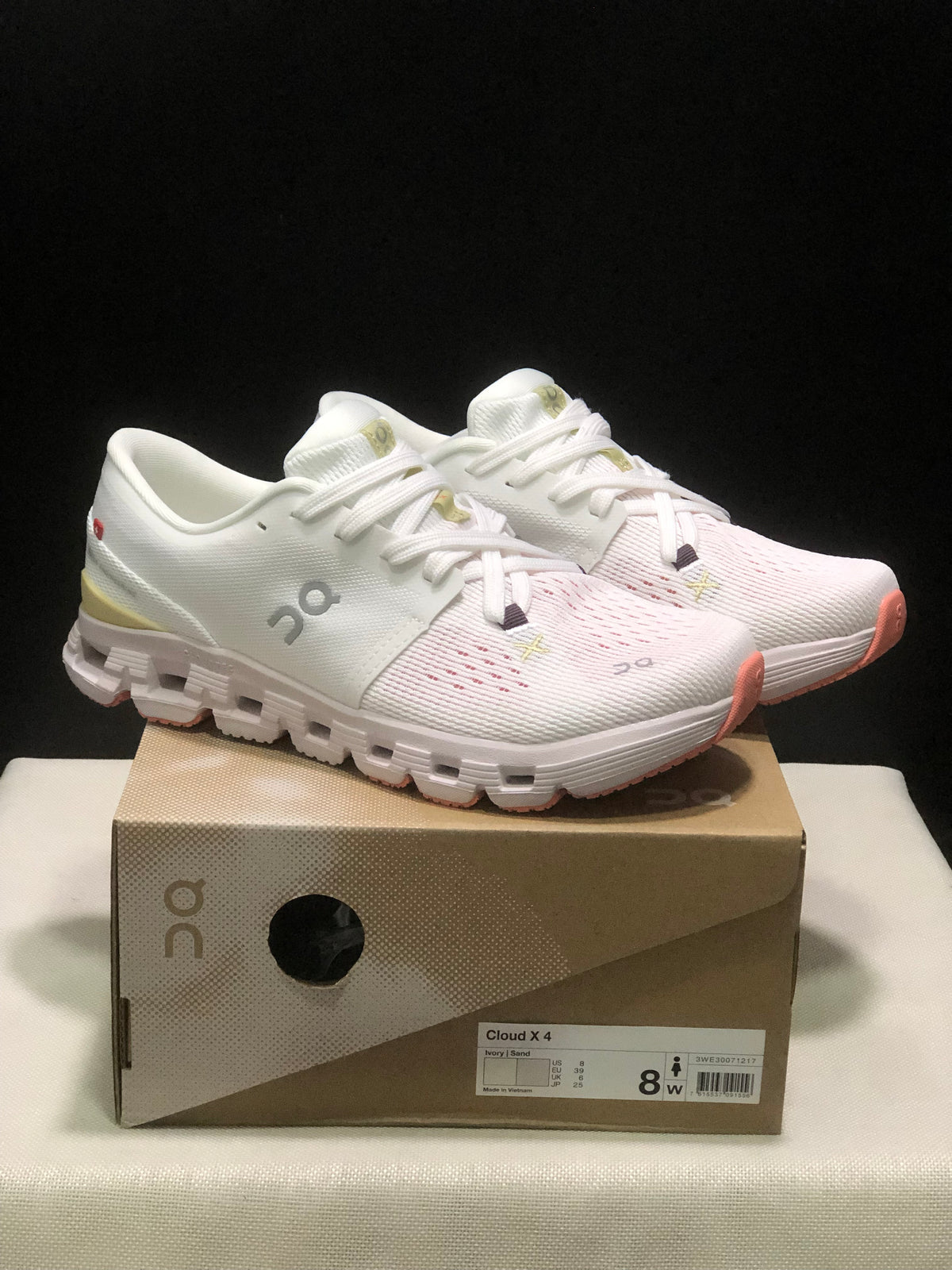 Pair of ivory and sand On Cloud X4 low-top running shoes displayed on a labeled shoe box.