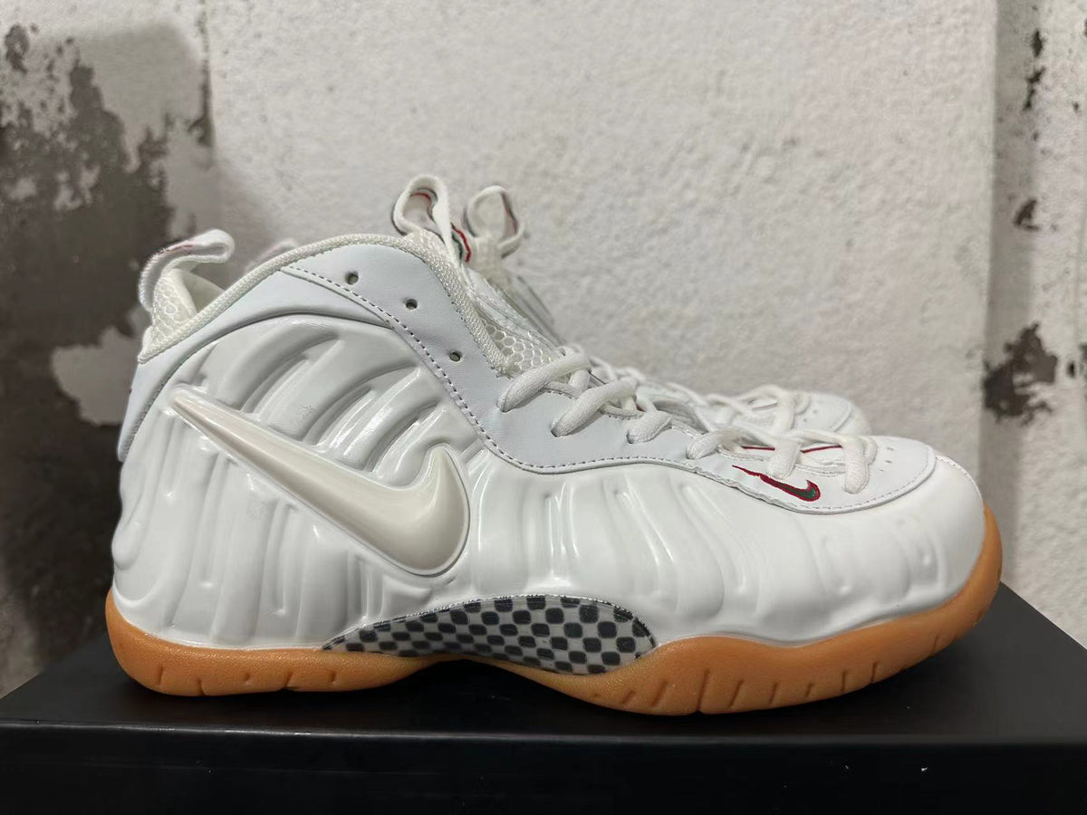 Side view of a white Nike Foamposite sneaker with sculpted panels, a raised Swoosh, checkerboard detail, and gum outsole