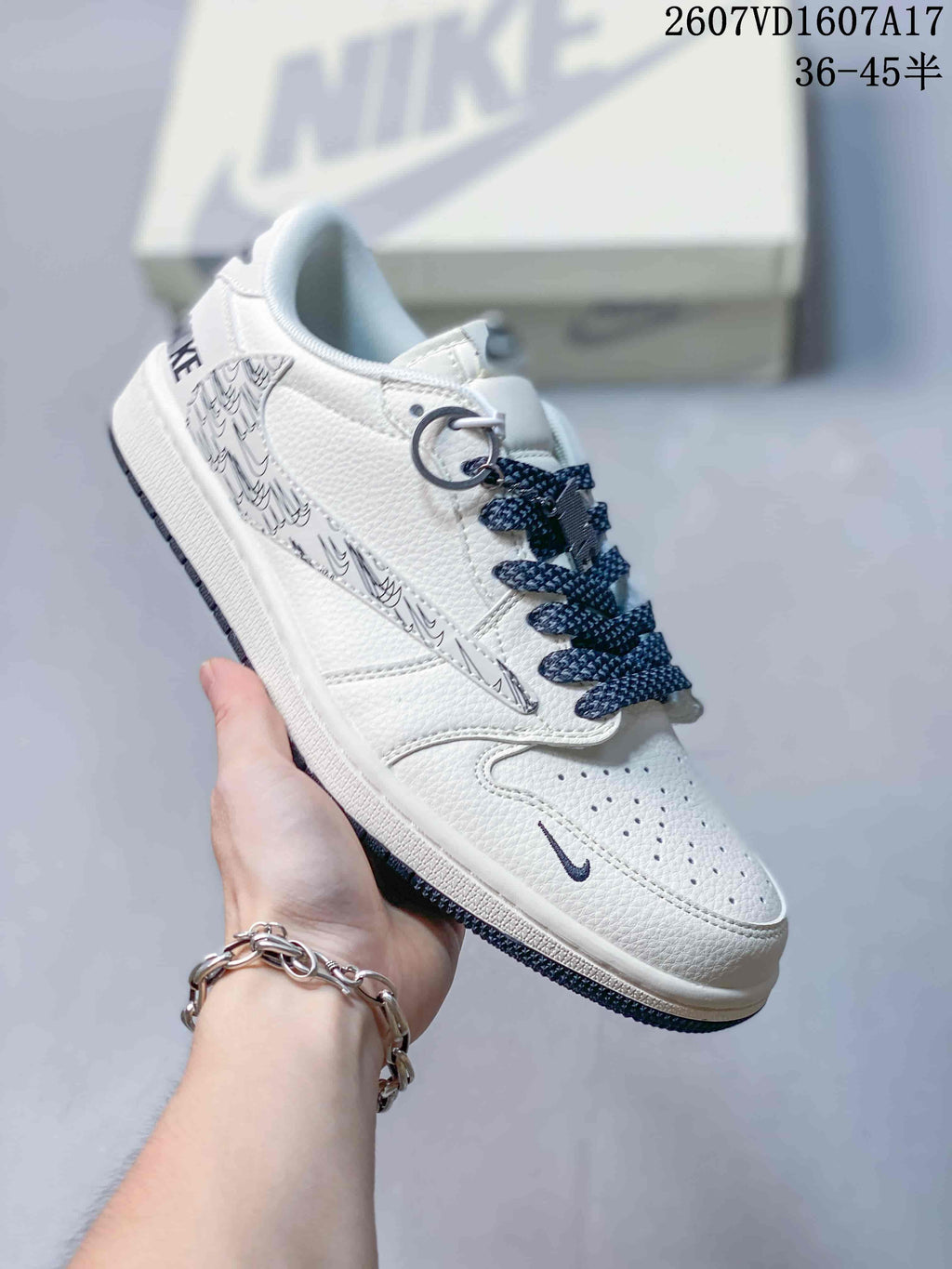 White low-top sneaker with monochrome graphic side panel, dark laces, metal lace accessories and a pale branded box behind it.