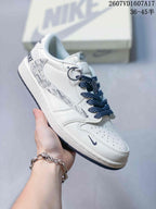 White low-top sneaker with monochrome graphic side panel, dark laces, metal lace accessories and a pale branded box behind it.