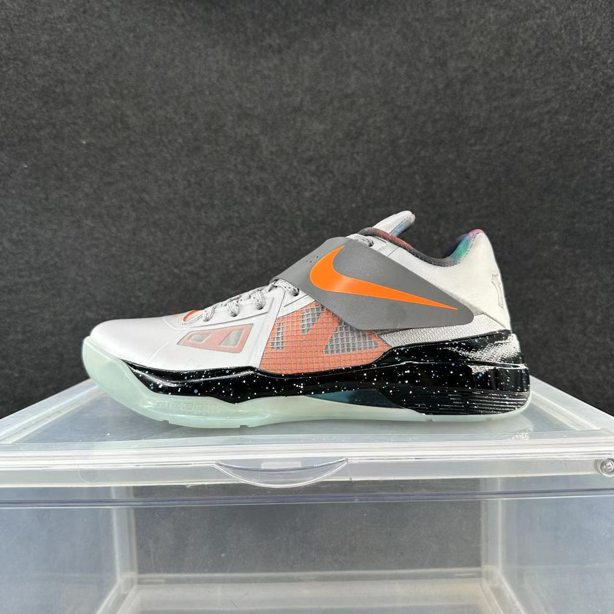 Side view of a white and gray Nike basketball sneaker with orange accents, a midfoot strap, black speckled midsole section, and translucent-looking outsole.