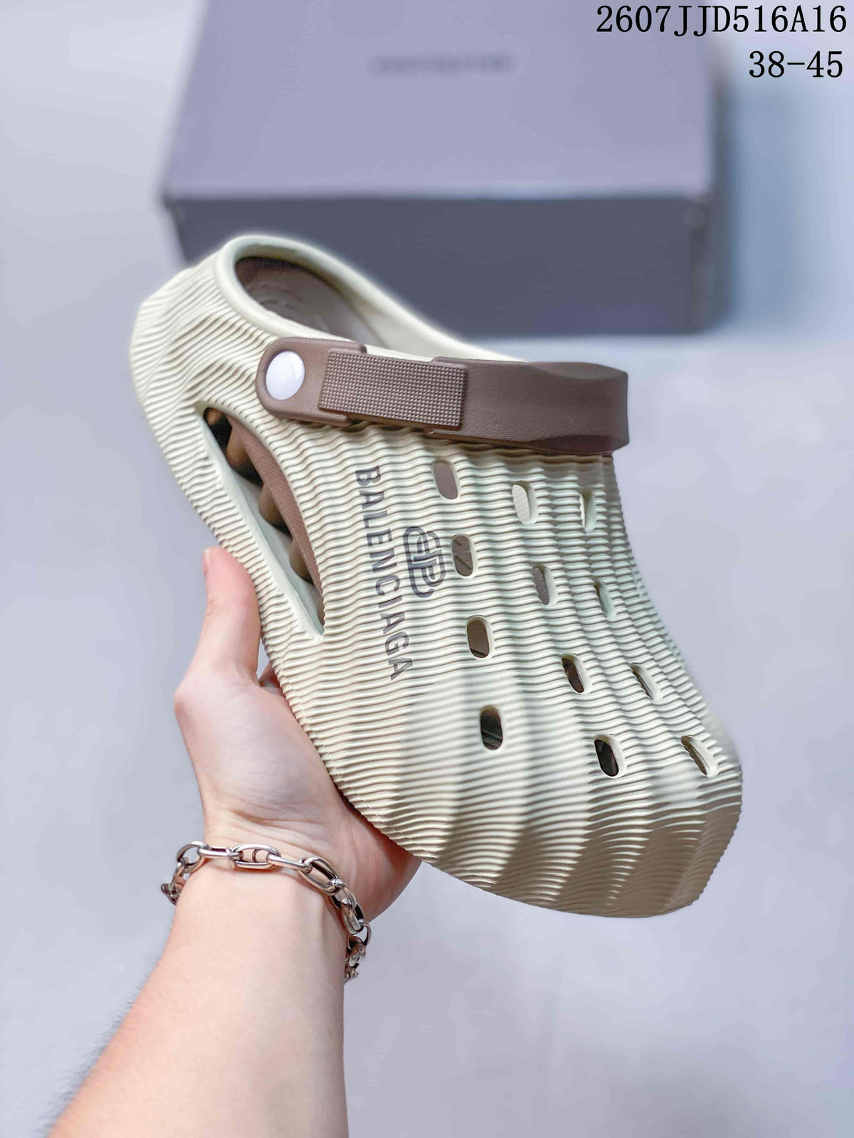 Angled side view of a cream clog slide showing the covered toe, ribbed construction, side branding, ventilation openings, and brown heel strap.