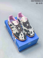 Pair of adidas Samba OG sneakers with a black-and-white cow-print upper, pink lining and gum outsole displayed on a blue shoebox.