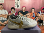 Side view of a light gray-white basketball shoe with gold-colored Swoosh, embossed geometric detailing, and a hanging charm.