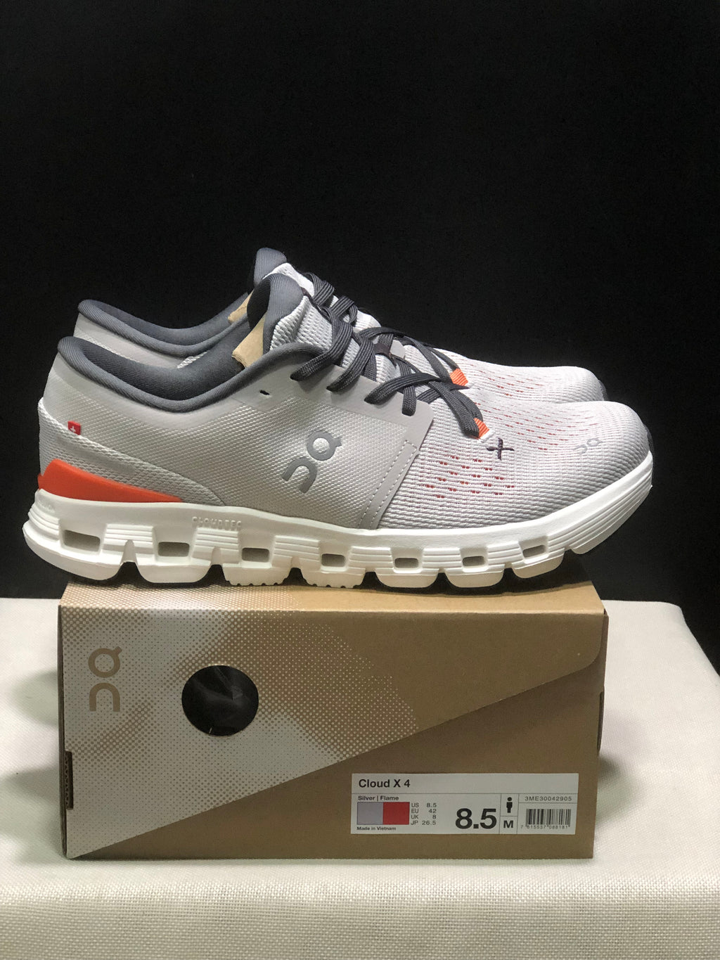 Side view of the silver and flame On Cloud X4 shoes showing the layered upper, orange heel accents, and white segmented outsole.