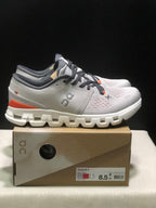 Side view of the silver and flame On Cloud X4 shoes showing the layered upper, orange heel accents, and white segmented outsole.