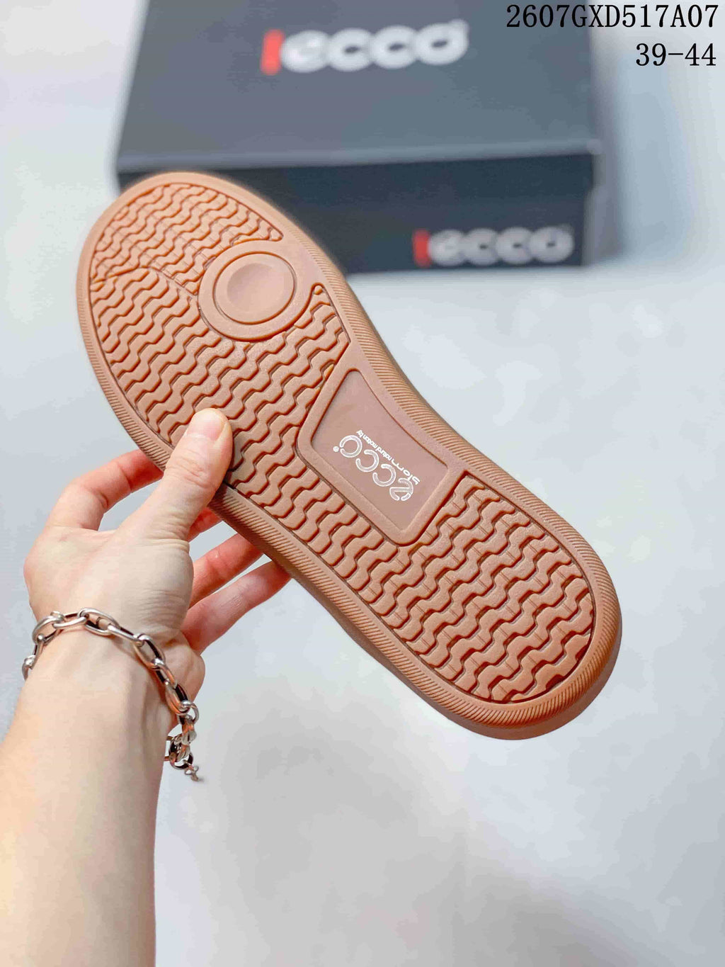 Bottom view of the sneaker showing the gum-colored rubber outsole with a repeating textured tread pattern; ECCO branding and size range 39-44 are visible in the background.