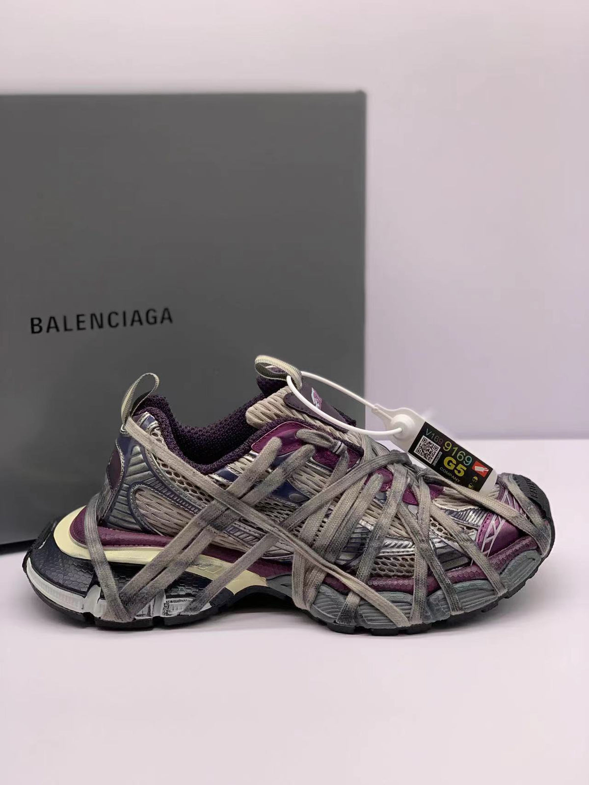 Side view of gray and purple Balenciaga 3XL sneakers with extended laces and a Balenciaga box in the background.