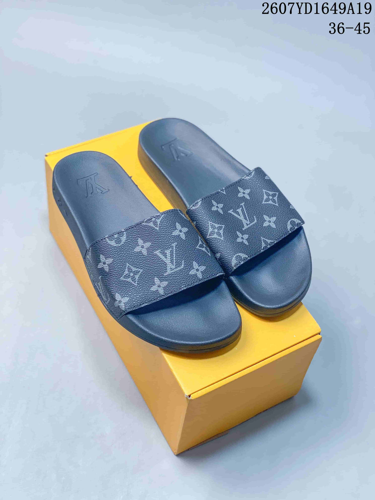 Pair of blue-gray monogram slide sandals displayed on a yellow box.