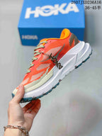 Side profile of the coral Hoka trail running shoe showing mesh panels, layered overlays, thick white sole, and dark heel detail.