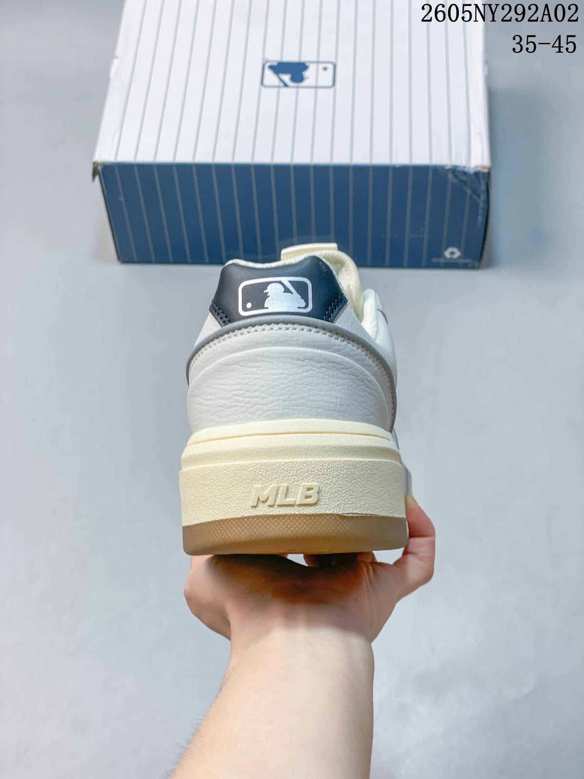 Rear view of a white sneaker showing the dark heel label, raised MLB lettering on the sole, and cream-toned heel section.