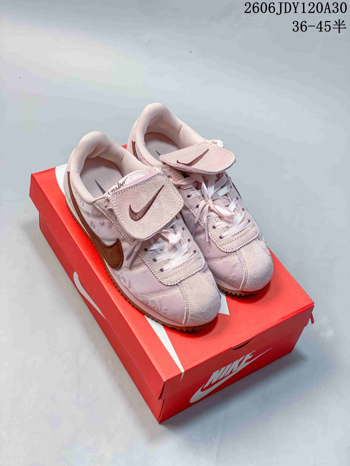 Pair of pale pink Nike Classic Cortez sneakers with brown accents and tongue flaps, shown on a red shoebox