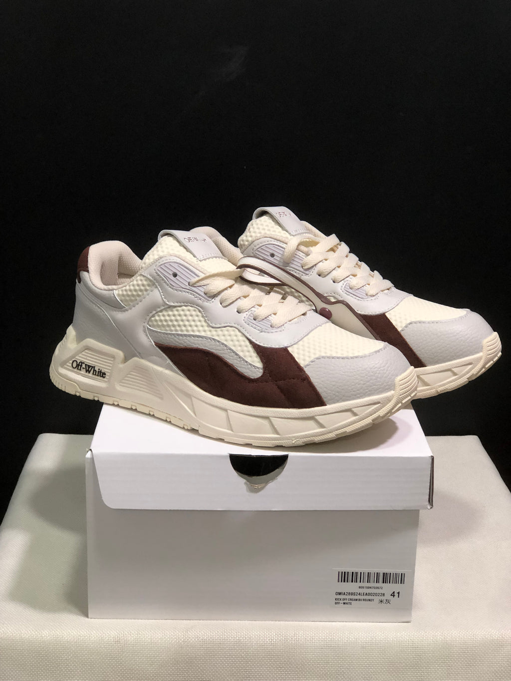Pair of white and brown low-top sneakers displayed on a white shoebox, showing the side profile and sculpted sole.