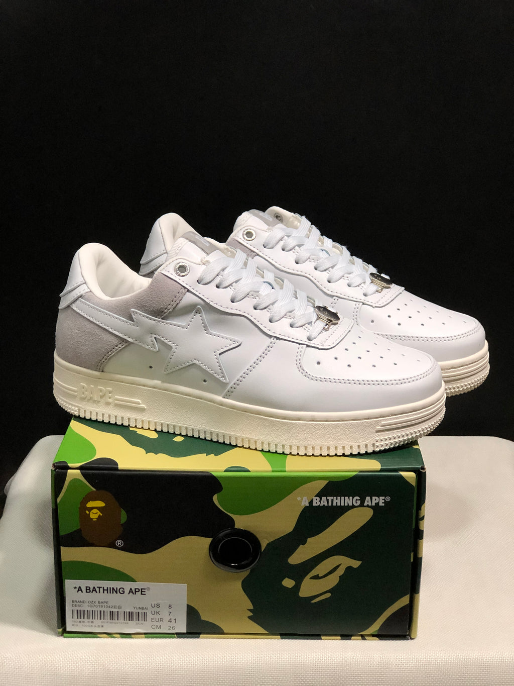 Pair of white BAPE Sta low-top sneakers with gray heel panels displayed on a camouflage shoebox.