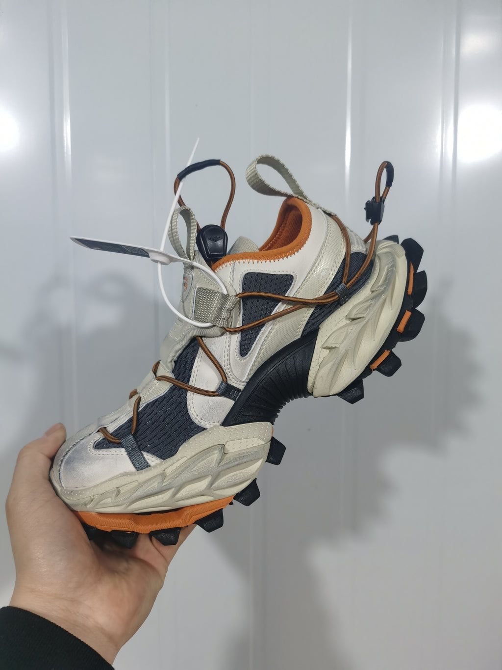 Side view of the beige, black, and orange sneaker highlighting the layered upper, orange trim, webbing details, and rugged outsole.