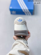 Rear view of the white sneaker showing the padded heel, blue trefoil-style branding, and dark outsole edge.