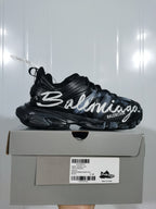 Side view of a black Balenciaga Track sneaker with white graffiti lettering displayed on its shoebox.