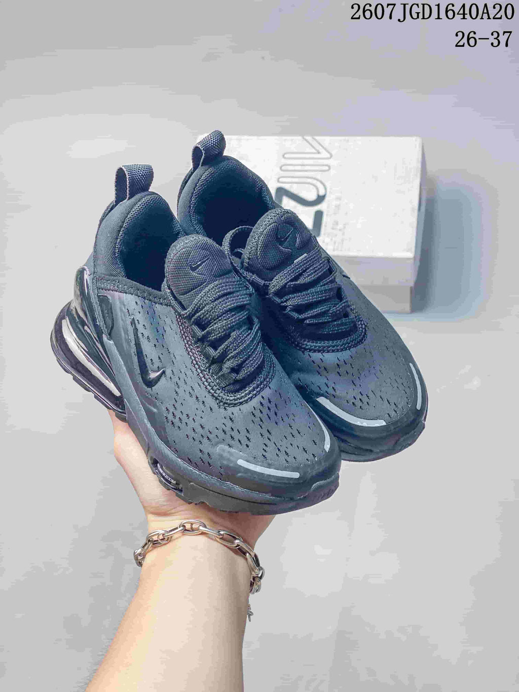 Pair of black Nike Air Max 270 children's sneakers held in front of a shoebox; overlay shows style text and the range 26–37.