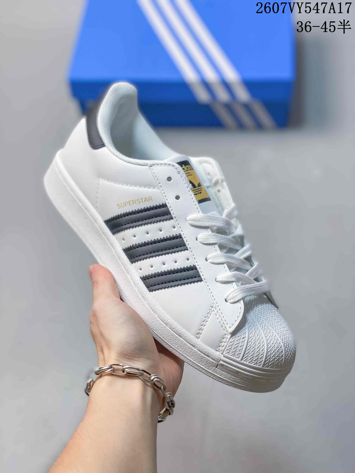 Angled side view of a white Adidas Superstar sneaker with black stripes and gold SUPERSTAR lettering.