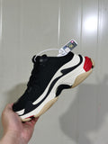 Side view of a black and white Balenciaga sneaker with red and beige sole accents, held against a light background.